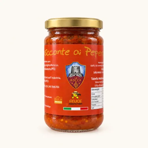 Glass jar of pepper sauce with a red-labelled package, held by a hand. In the background you can see vegetables, a chopping board and other kitchen tools.